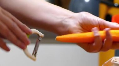 Temiz beyaz arka planda metal kabuklu taze, canlı turuncu bir havucu soyan ellerin yakın çekimi. Sağlıklı beslenme, yemek pişirme ve yemek hazırlama kavramı.