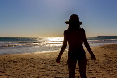 Chica joven con sombrero mirando al mar en un atardecer. Seçici odaklanma. Solunum konsepti. Arka ışıklandırma..