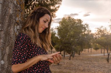 Sonbaharda bir ağaca yaslanıp ellerine bakan genç bir kadın. Seçici odak.