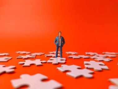 Tek bir yapboz parçasının üzerinde duran minyatür insan biblosu, turuncu bir arka planda dağınık parçalarla çevrili..