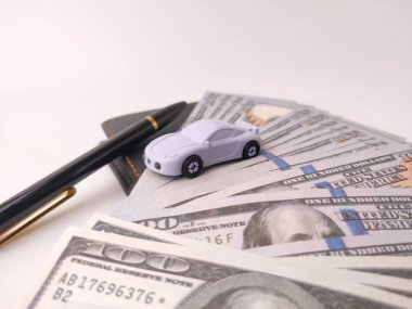 Bu resim, ABD dolarlarının üzerinde duran küçük bir oyuncak araba modelini ve muhtemelen araçlarla ilgili finansal kavramları sembolize eden bir kalemi gösteriyor..