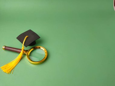 Görüntü yeşil arka planda minyatür kare akademik şapkanın yanında büyüteç gösteriyor.