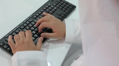 Kandura 'da masa başında oturan ve bilgisayar klavyesinde yazan Arap bir adam. Geleneksel emirlik elbiseli erkek Arap yönetici daktilo kullanıyor.