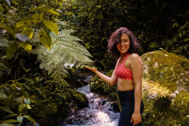 Egzotik bir kadın gülüyor ve ormanda büyük bir bitkiye dokunuyor.