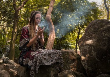 Rastalı beyaz kadın doğada tütsü yakıyor.