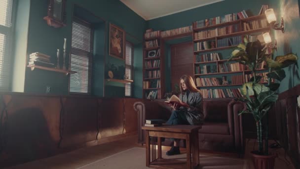 Giovane donna caucasica attraente sfoglia le pagine e fa conoscenza con il contenuto del libro mentre si siede sul divano in biblioteca — Video Stock