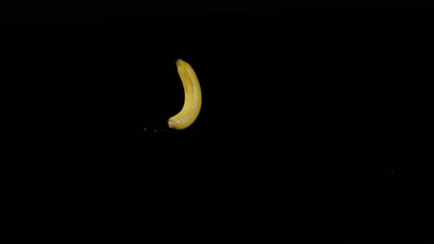 Υγρή φρέσκια μπανάνα πετάει πάνω και περιστρέφεται με πιτσιλιές νερού σε μαύρο φόντο σε αργή κίνηση σουτ, νερό πέφτει στα φρούτα — Αρχείο Βίντεο