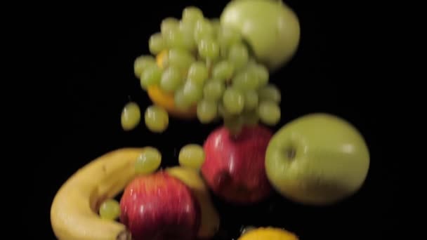 Cerca de las uvas frescas, manzanas, plátanos, limón y peras sacudidas y girando sobre un fondo negro en cámara lenta, gotas de agua en las frutas — Vídeo de stock