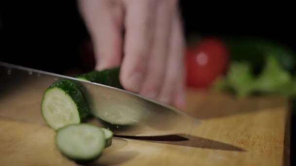 Närbild på mens händer skära gurka med en kniv i bitar på skärbräda på en svart bakgrund — Stockvideo