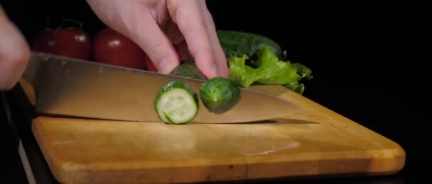 Mens hands cut fresh juicy cucumber with a knife into pieces on cutting board on a black background — Stock Video