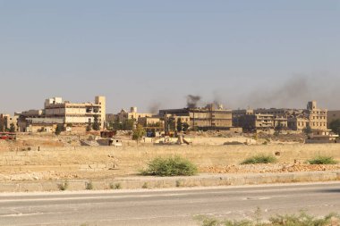 Savaştan zarar görmüş bir şehirde fabrika bacasından yoğun duman yükseliyor. Sahne, yoğun kirlilik, çevre standartlarının eksikliği ve kentsel sanayi üzerindeki çatışmaların yıkıcı etkilerini vurguluyor..