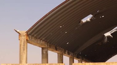 Suriye 'nin Halep cephesindeki yok edilmiş bir depo. Metal kemerli çatı, muhtemelen havan mermisinin neden olduğu patlama hasarını gösteriyor. Sahne, savaş yıkımını ve kentsel dayanıklılığı yansıtıyor..