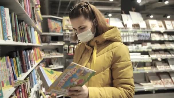 Mulher em máscara médica protetora escolhe livros no supermercado durante a epidemia de coronavírus covid-19. Compras durante a quarentena, coronavírus COVID-19 pandemia. — Vídeo de Stock