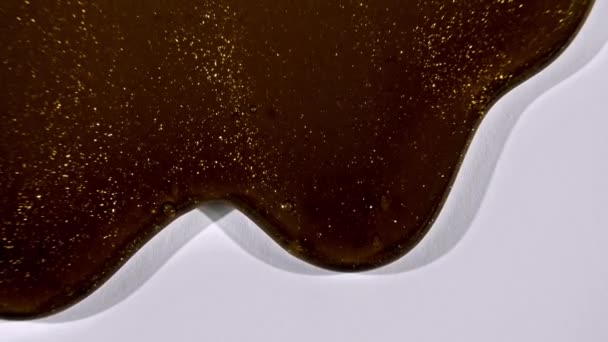 Gel cosmético marrón fluido dorado intercalado fluyendo con hacia abajo sobre una superficie blanca. Macro Shot — Vídeos de Stock