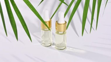 Doğal kozmetik malzemeli damlatan cam şişe, gerekli yağ. Beyaz arka planda gölgeli tropikal palmiye yapraklarının hareketi.