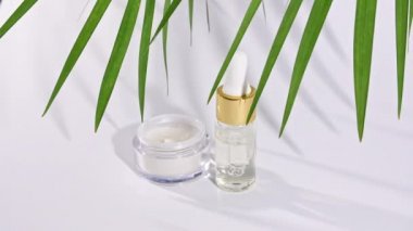 Doğal kozmetik malzemeli damlatan cam şişe, gerekli yağ. Beyaz yüz kremi kavanozu. Deri ve vücut bakımı. Beyaz arka planda gölgeli tropikal palmiye yapraklarının hareketi.