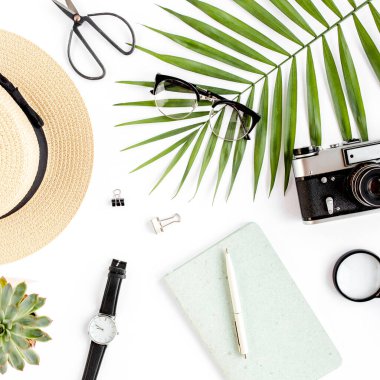 Üst kat ofis masası. Hasır şapka, gözlük, retro kamera, not defteri ve beyaz arka planda tropikal palmiye yaprağı olan çalışma alanı. Düz yatış, üst görünüm.