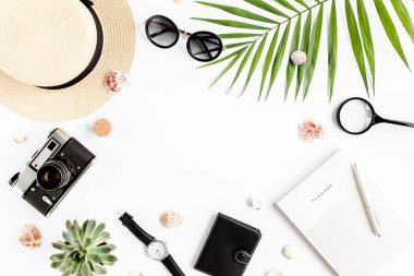 Üst kat ofis masası. Hasır şapka, gözlük, retro kamera, not defteri ve beyaz arka planda tropikal palmiye yaprağı olan çalışma alanı. Düz yatış, üst görünüm.