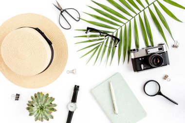 Üst kat ofis masası. Hasır şapka, gözlük, retro kamera, not defteri ve beyaz arka planda tropikal palmiye yaprağı olan çalışma alanı. Düz yatış, üst görünüm.