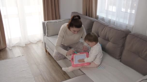 Fürsorgliche junge Mutter oder Kindermädchen, die ihrer süßen Tochter hilft, dem Baby beizubringen, ein Bild mit Bleistiften zu zeichnen, auf dem Sofa im Wohnzimmer zu liegen, zu Hause Kreativität zu erlernen — Stockvideo