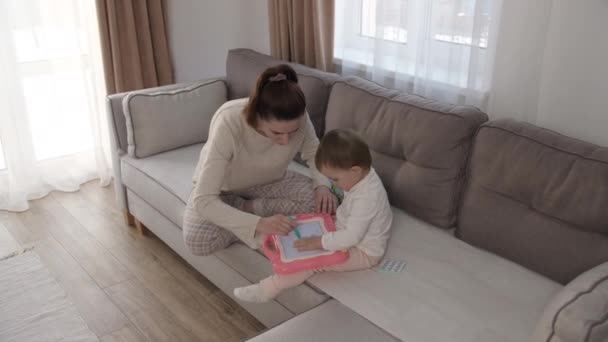 Cuidar a la joven madre o niñera ayudando a su linda hija a enseñar al bebé, dibujar un cuadro con lápices, acostado en el sofá de la sala de estar, aprender creatividad en casa — Vídeos de Stock