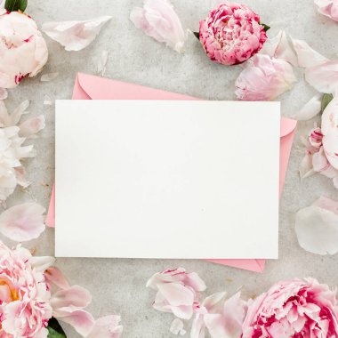 Sahte davetiye, boş kağıt tebrik kartı, pembe zarf ve gri taş masada şakayıklar. Çiçek arkaplanı. Düz yatış, üst görünüm.