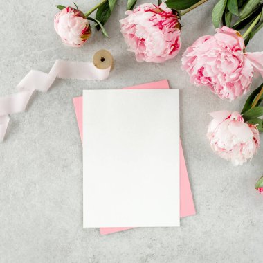 Sahte davetiye, boş kağıt tebrik kartı, pembe zarf ve gri taş masada şakayıklar. Çiçek arkaplanı. Düz yatış, üst görünüm.