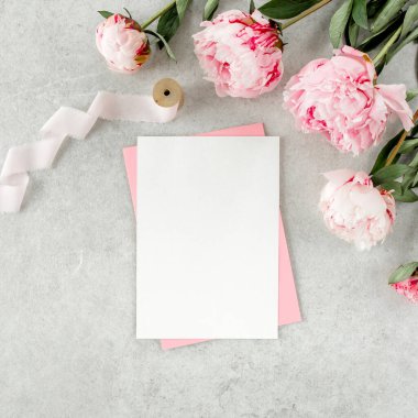 Sahte davetiye, boş kağıt tebrik kartı, pembe zarf ve gri taş masada şakayıklar. Çiçek arkaplanı. Düz yatış, üst görünüm.