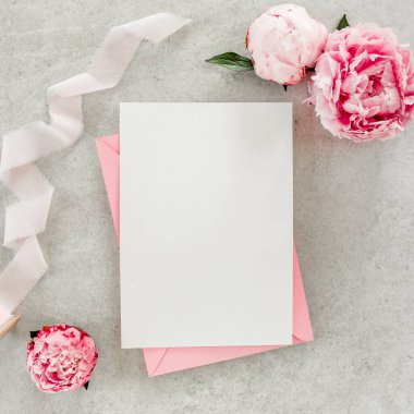 Sahte davetiye, boş kağıt tebrik kartı, pembe zarf ve gri taş masada şakayıklar. Çiçek arkaplanı. Düz yatış, üst görünüm.