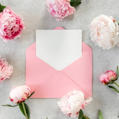 Sahte davetiye, boş kağıt tebrik kartı, pembe zarf ve gri taş masada şakayıklar. Çiçek arkaplanı. Düz yatış, üst görünüm.