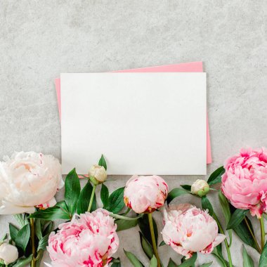 Sahte davetiye, boş kağıt tebrik kartı, pembe zarf ve gri taş masada şakayıklar. Çiçek arkaplanı. Düz yatış, üst görünüm.
