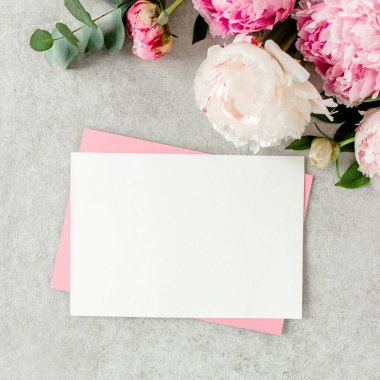 Sahte davetiye, boş kağıt tebrik kartı, pembe zarf ve gri taş masada şakayıklar. Çiçek arkaplanı. Düz yatış, üst görünüm.