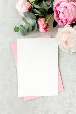 Sahte davetiye, boş kağıt tebrik kartı, pembe zarf ve gri taş masada şakayıklar. Çiçek arkaplanı. Düz yatış, üst görünüm.