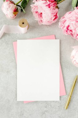 Sahte davetiye, boş kağıt tebrik kartı, pembe zarf ve gri taş masada şakayıklar. Çiçek arkaplanı. Düz yatış, üst görünüm.