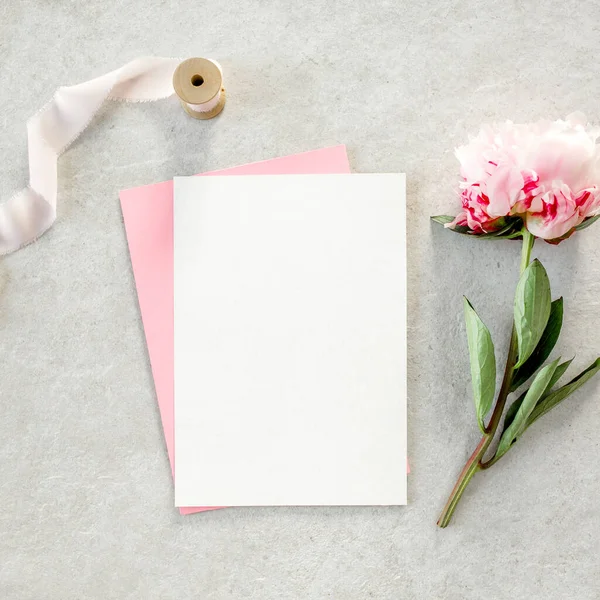 Sahte davetiye, boş kağıt tebrik kartı, pembe zarf ve gri taş masada şakayıklar. Çiçek arkaplanı. Düz yatış, üst görünüm.