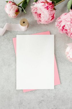 Sahte davetiye, boş kağıt tebrik kartı, pembe zarf ve gri taş masada şakayıklar. Çiçek arkaplanı. Düz yatış, üst görünüm.