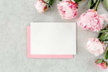 Sahte davetiye, boş kağıt tebrik kartı, pembe zarf ve gri taş masada şakayıklar. Çiçek arkaplanı. Düz yatış, üst görünüm.