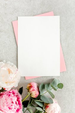 Sahte davetiye, boş kağıt tebrik kartı, pembe zarf ve gri taş masada şakayıklar. Çiçek arkaplanı. Düz yatış, üst görünüm.