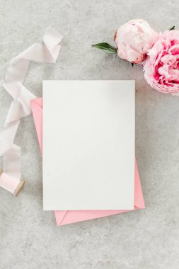 Sahte davetiye, boş kağıt tebrik kartı, pembe zarf ve gri taş masada şakayıklar. Çiçek arkaplanı. Düz yatış, üst görünüm.