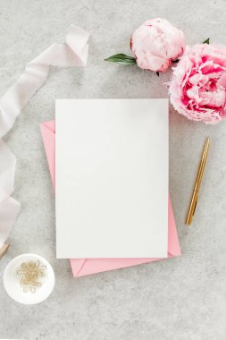 Sahte davetiye, boş kağıt tebrik kartı, pembe zarf ve gri taş masada şakayıklar. Çiçek arkaplanı. Düz yatış, üst görünüm.