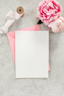 Sahte davetiye, boş kağıt tebrik kartı, pembe zarf ve gri taş masada şakayıklar. Çiçek arkaplanı. Düz yatış, üst görünüm.