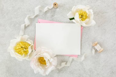 Sahte davetiye, boş kağıt tebrik kartı, pembe zarf ve gri taş masada şakayıklar. Çiçek arkaplanı. Düz yatış, üst görünüm.