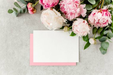 Sahte davetiye, boş kağıt tebrik kartı, pembe zarf ve gri taş masada şakayıklar. Çiçek arkaplanı. Düz yatış, üst görünüm.