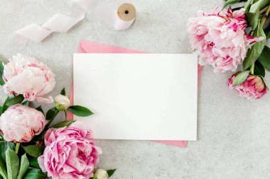 Sahte davetiye, boş kağıt tebrik kartı, pembe zarf ve gri taş masada şakayıklar. Çiçek arkaplanı. Düz yatış, üst görünüm.