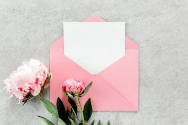 Sahte davetiye, boş kağıt tebrik kartı, pembe zarf ve gri taş masada şakayıklar. Çiçek arkaplanı. Düz yatış, üst görünüm.