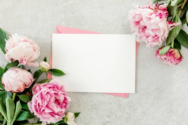 Sahte davetiye, boş kağıt tebrik kartı, pembe zarf ve gri taş masada şakayıklar. Çiçek arkaplanı. Düz yatış, üst görünüm.