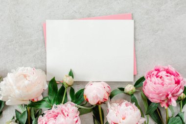 Sahte davetiye, boş kağıt tebrik kartı, pembe zarf ve gri taş masada şakayıklar. Çiçek arkaplanı. Düz yatış, üst görünüm.