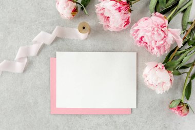 Sahte davetiye, boş kağıt tebrik kartı, pembe zarf ve gri taş masada şakayıklar. Çiçek arkaplanı. Düz yatış, üst görünüm.