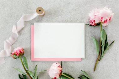 Sahte davetiye, boş kağıt tebrik kartı, pembe zarf ve gri taş masada şakayıklar. Çiçek arkaplanı. Düz yatış, üst görünüm.