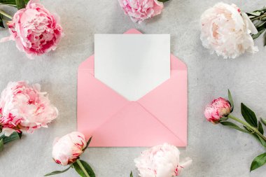 Sahte davetiye, boş kağıt tebrik kartı, pembe zarf ve gri taş masada şakayıklar. Çiçek arkaplanı. Düz yatış, üst görünüm.